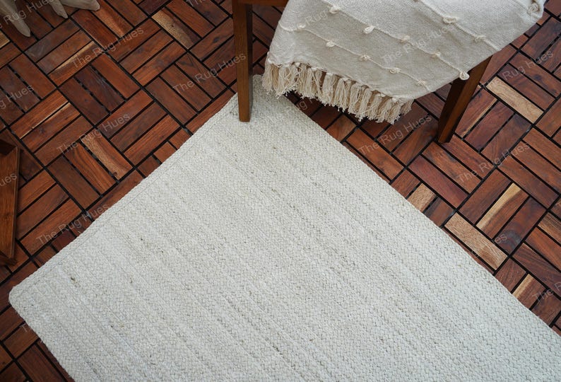 May include: A white woven rug with a textured surface. The rug is placed on a wooden floor with a herringbone pattern.