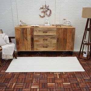May include: A white rectangular jute rug with a natural woven texture, placed on a wooden floor with a tiled pattern. The rug is in front of a wooden cabinet with three drawers and two doors. The cabinet has a natural wood finish and black metal legs.