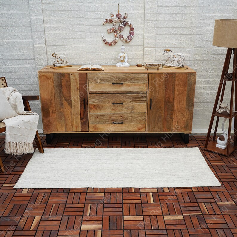 May include: A white rectangular jute rug with a natural woven texture, placed on a wooden floor with a tiled pattern. The rug is in front of a wooden cabinet with three drawers and two doors. The cabinet has a natural wood finish and black metal legs.