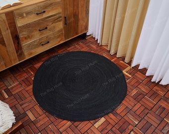 Puede incluir: Una alfombra redonda de yute trenzado negra con un patrón circular. La alfombra está sobre un suelo de madera con un patrón de azulejos.
