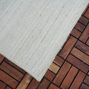 May include: A white woven rug with a textured surface. The rug is placed on a dark brown wood floor.