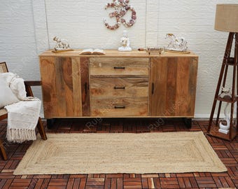 May include: A natural jute rug with a woven border in a light brown color. The rug is placed in front of a wooden cabinet with three drawers and two doors. The cabinet is sitting on a brown tiled floor.