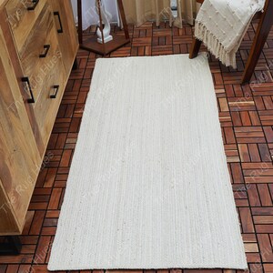 May include: A white, woven, rectangular rug with a subtle texture. The rug is laid out on a wooden floor.