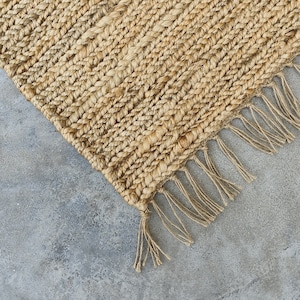 Puede incluir: Primer plano de una alfombra de fibra natural con borde de flecos. La alfombra está hecha de yute tejido y tiene un color marrón claro. La alfombra está sobre un suelo de hormigón.