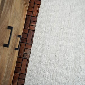 May include: A white woven rug with a textured surface. The rug is placed on top of a wooden floor with a pattern of brown tiles.