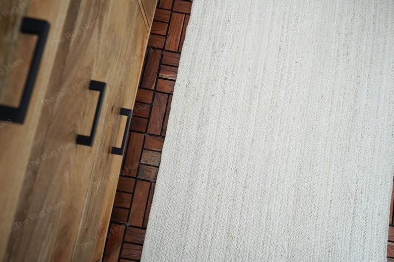 May include: A white woven rug with a textured surface. The rug is placed on top of a wooden floor with a pattern of brown tiles.