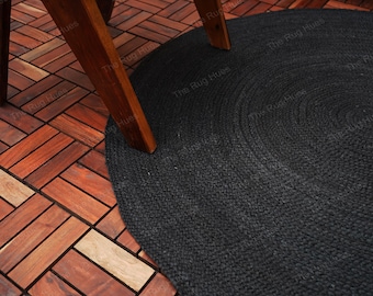 Puede incluir: Una alfombra redonda de yute negra con un patrón tejido. La alfombra está colocada sobre un suelo de madera con una pata de silla apoyada en el borde de la alfombra.