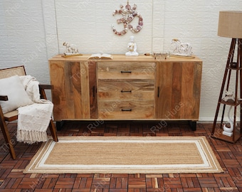 May include: A natural fiber rug with a white border, placed in front of a wooden cabinet with three drawers. The cabinet is in a room with a tiled floor and a white wall with a decorative Om symbol made of seashells.