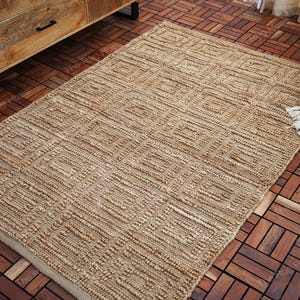 Puede incluir: Una alfombra de fibra natural con un patrón geométrico de color marrón claro. La alfombra está hecha de yute y tiene una textura tejida. Está colocada sobre un suelo de madera.
