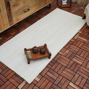 May include: A white rectangular jute rug with a natural woven texture. The rug is placed on a wooden floor with a herringbone pattern. Two small ceramic containers are on a wooden tray.