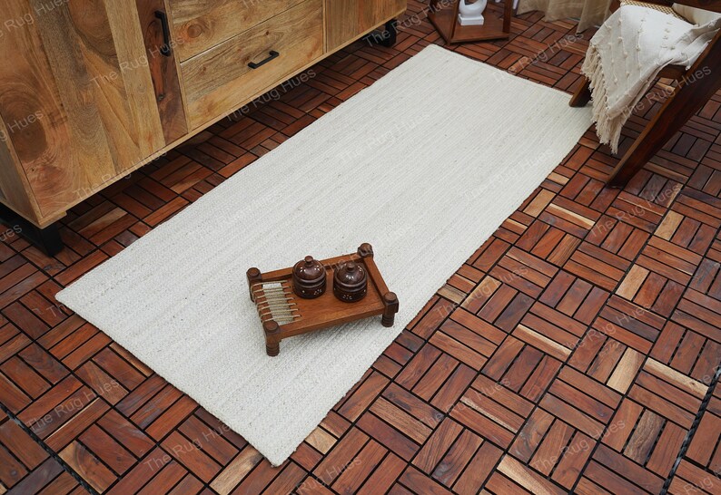 May include: A white rectangular jute rug with a natural woven texture. The rug is placed on a wooden floor with a herringbone pattern. Two small ceramic containers are on a wooden tray.