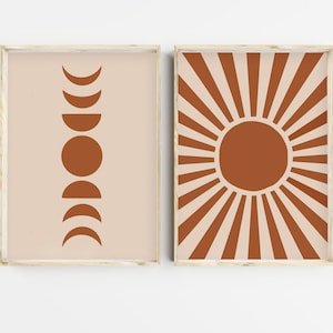Puede incluir: Dos impresiones enmarcadas con un diseño minimalista. La impresión de la izquierda presenta una luna marrón en varias fases sobre un fondo beige claro. La impresión de la derecha presenta un sol marrón con rayos que emanan de él sobre un fondo beige claro.