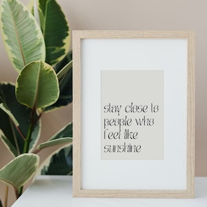 Puede incluir: Una impresión enmarcada con fondo beige y texto negro que dice "Quédate cerca de las personas que te hacen sentir como el sol".
