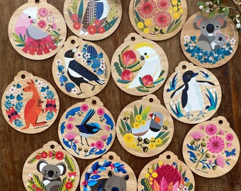 May include: Twelve round wooden ornaments featuring various Australian animals and plants. The ornaments are decorated with colorful illustrations of native animals like a kookaburra, a penguin, a wombat, a koala, and a kangaroo. The ornaments also feature native plants like banksia and wattle.