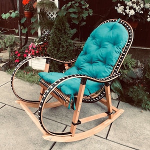 Könnte beinhalten: Ein Holz-Schaukelstuhl mit einem türkisfarbenen Kissen. Der Stuhl hat einen schwarzen und weißen Besatz an den Rändern. Der Stuhl steht auf einer Terrasse.