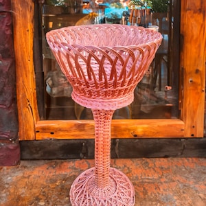 May include: A pink wicker basket with a tall stand. The basket is woven with a delicate pattern and has a wide, open top.