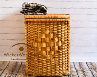 May include: A woven wicker basket with a plaid fabric folded on top. The basket is brown and has a rectangular shape. The basket is sitting on a wooden floor in front of a white brick wall.