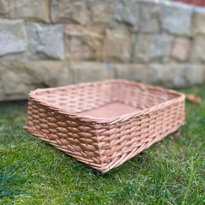 Può includere: Un cesto rettangolare in vimini intrecciato, appoggiato su un letto di erba verde. Il cesto è di colore marrone chiaro e ha un aspetto naturale e rustico.
