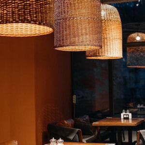 Könnte beinhalten: Ein Restaurant-Interieur mit Holztischen und -stühlen. Die Tische sind mit Salz- und Pfefferstreuern gedeckt. Das Restaurant hat eine warme, einladende Atmosphäre mit geflochtenen Pendelleuchten, die von der Decke hängen.