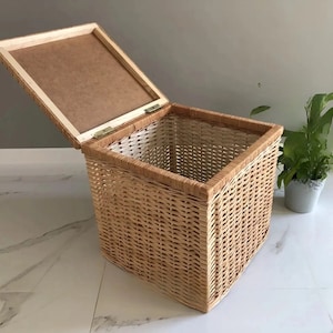 Pode incluir: Uma cesta de armazenamento de vime tecida com uma tampa de madeira. A cesta é de cor castanho claro e tem uma forma quadrada. A tampa está aberta e a cesta está sentada num chão de azulejos brancos.