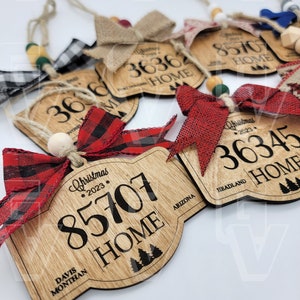 Puede incluir: Un conjunto de cinco adornos navideños de madera con cinta de cuadros rojos y negros. Cada adorno tiene un diseño en forma de casa con un código postal, un estado y la palabra "Home".