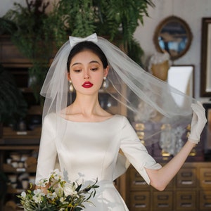 Könnte beinhalten: Eine Braut in einem weißen Satin-Hochzeitskleid mit einem weißen Schleier und einer weißen Schleife. Sie hält einen Strauß weißer Blumen.