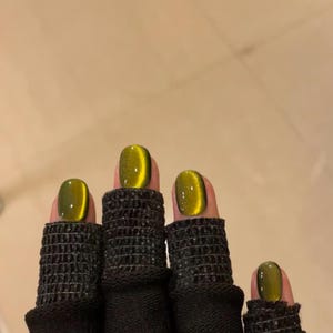 May include: Close-up of four fingers wearing black finger cots with green and gold cat eye nail polish.
