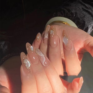 May include: Close-up of a hand with long, almond-shaped nails. The nails are painted with a sheer nude polish and decorated with gold glitter and white swirls.