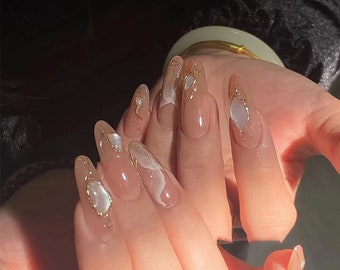 May include: Close-up of a hand with long, almond-shaped nails. The nails are painted with a sheer nude polish and decorated with gold glitter and white swirls.