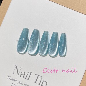 May include: Six light blue glitter press-on nails with a shimmery finish. The nails are arranged in a row on a white surface. The text "Nail Tip" and "Ccstr nail" is visible in the image.