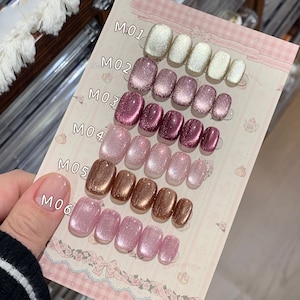Könnte beinhalten: Sechs Reihen mit aufklebbaren Nägeln mit Glitzer-Finish in verschiedenen Farben, darunter Weiß, Rosa, Gold und Burgunderrot. Jede Reihe ist mit einer Nummer von M01 bis M06 gekennzeichnet.