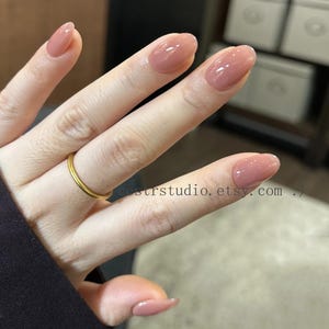 Puede incluir: Primer plano de una mano con uñas ovaladas pintadas en un suave tono rosa apagado. Las uñas tienen un acabado brillante. Un fino anillo dorado adorna el dedo anular. La URL de la tienda Etsy es visible.