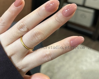 Puede incluir: Primer plano de una mano con uñas ovaladas pintadas en un suave tono rosa apagado. Las uñas tienen un acabado brillante. Un fino anillo dorado adorna el dedo anular. La URL de la tienda Etsy es visible.