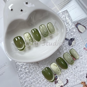Op de afbeelding: Set van tien groene en witte press-on nagels met een bladerenontwerp. De nagels zijn gerangschikt in een witte kom.