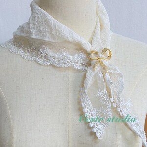 May include: White lace scarf with a gold bow brooch featuring a pearl accent. The scarf is draped over a white mannequin.