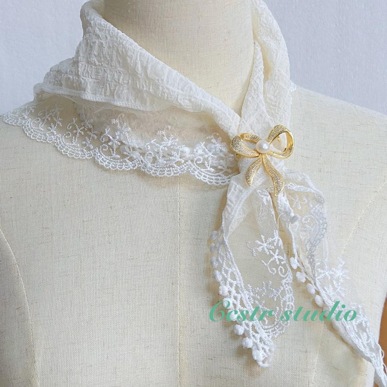 May include: White lace scarf with a gold bow brooch featuring a pearl accent. The scarf is draped over a white mannequin.