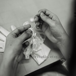 May include: A pair of white gloves featuring delicate lace trim and small buttons being hand-stitched with a needle and thread. The fabric is smooth and light, with intricate floral patterns on the lace edges. Text reads cestrstudio.etsy.com :)