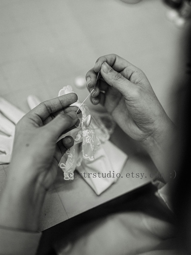 May include: A pair of white gloves featuring delicate lace trim and small buttons being hand-stitched with a needle and thread. The fabric is smooth and light, with intricate floral patterns on the lace edges. Text reads cestrstudio.etsy.com :)