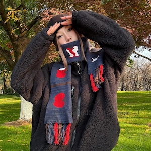 May include: A person wearing a black fleece jacket, a black beret, and a patterned scarf. The scarf is navy blue with red heart-shaped designs and fringed ends. The background features trees with colorful autumn leaves.