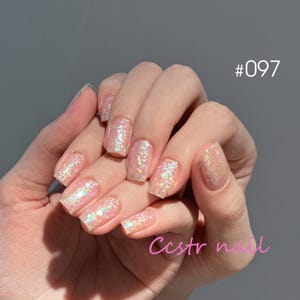 Könnte beinhalten: Nahaufnahme einer Hand mit kurzen, quadratischen Nägeln, die mit einem hellrosa Glitzerlack lackiert sind. Die Nägel sind mit schimmerndem Glitzer verziert. Das Bild ist mit #097 und "ccstr nail" beschriftet.