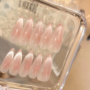 May include: Set of ten pink and white false nails with a shimmery finish. The nails are arranged in two rows in a clear plastic container.