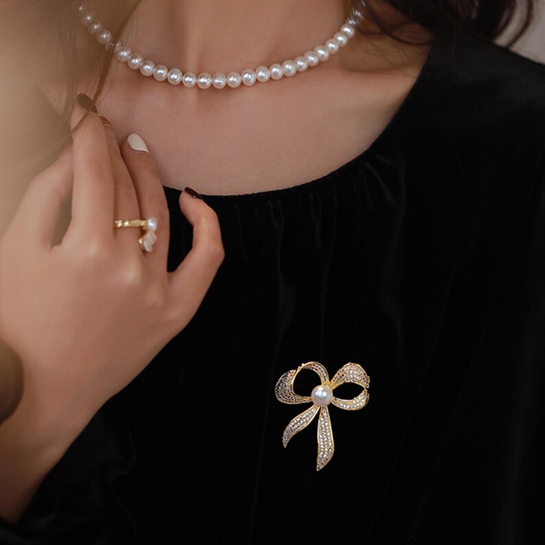 May include: A gold bow brooch with a pearl in the center, worn on a black velvet dress. The brooch is adorned with sparkling rhinestones.