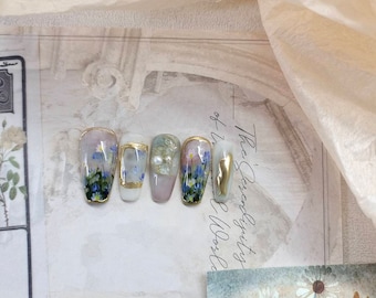 May include: Five nude-colored artificial nails with a floral design. Each nail has a gold accent and a pearl embellishment. The nails are arranged in a row on a white surface.