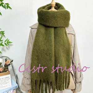 May include: A soft, green fuzzy scarf with long fringe tassels. The scarf is draped over a mannequin and a patterned fabric background.