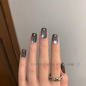 Op de afbeelding: Close-up van een hand met donkergrijze, vierkante nagels. Elke nagel heeft een klein, zilverkleurig sterontwerp. Een zilveren ring met een geometrisch patroon zit om de ringvinger. De tekst "ccstrstudio.etsy.com :)" is zichtbaar.
