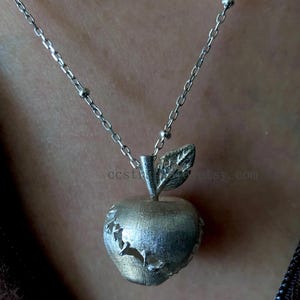 Puede incluir: Un collar con colgante de manzana plateada. La manzana tiene una superficie texturizada con diseños recortados. La cadena del collar es delicada, con pequeñas cuentas intercaladas a lo largo de su longitud. El colgante cuelga de la cadena.