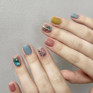Puede incluir: Primer plano de una mano con uñas cortas y cuadradas pintadas de varios colores. Las uñas están decoradas con purpurina y pedrería. Los colores incluyen azul claro, amarillo, rosa y un azul claro con una pedrería turquesa.