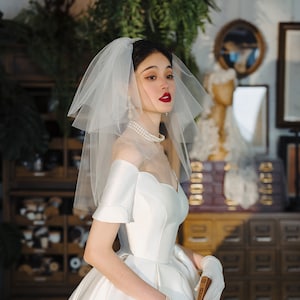 Puede incluir: Un vestido de novia blanco con escote de corazón y mangas cortas. El vestido tiene una falda completa y se combina con un velo blanco y un collar de perlas.