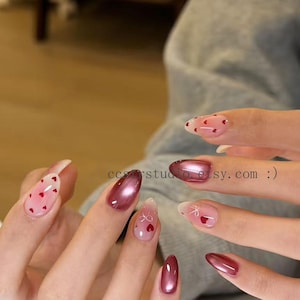 May include: A set of almond-shaped artificial nails in various designs. Some nails are a metallic burgundy, while others are clear with red heart and bow patterns. The text "ccrstudio.etsy.com :)" is visible.