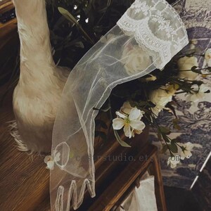 May include: A sheer white tulle glove featuring an intricate floral lace trim at the wrist. The glove is crafted from a delicate, semi-transparent mesh fabric and includes a circular cutout detail on the back of the hand, extending to the fingers.